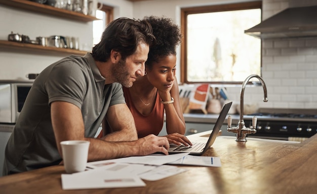 A young couple smiling and reviewing financial documents together at their kitchen table, laptop open in front of them, depicting financial planning and communication.