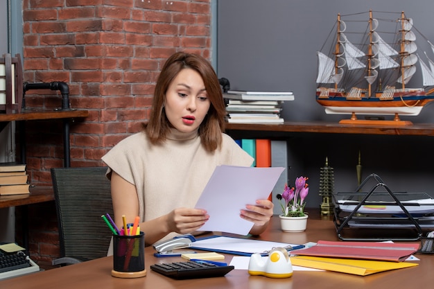 A woman is sitting at her desk, reviewing financial documents with a focused expression. She is using a laptop and calculator, with sticky notes and a cup of coffee nearby. The scene conveys a sense of diligence and financial awareness.