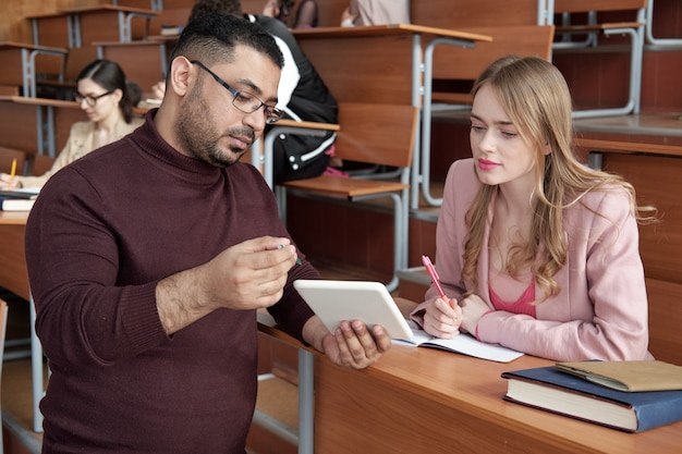 A mentor teacher observes a new teacher in a classroom, taking notes and providing constructive feedback. The new teacher is interacting with students.